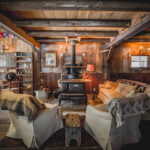 Rustic cozy cabin living room with cabincore vibes, woodstove, antiques, string lights, armchairs, and wooden ceiling beams.