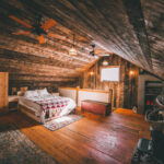 Cozy loft bedroom in rustic tiny cabin with wide wooden floorboards, pendant lighting fixtures, ceiling fans, and area rugs.