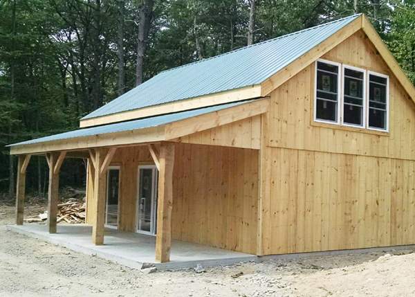 Rustic timber frame cabin with mortise and tenon post and beam construction, green roof, roof overhang over concrete slab.