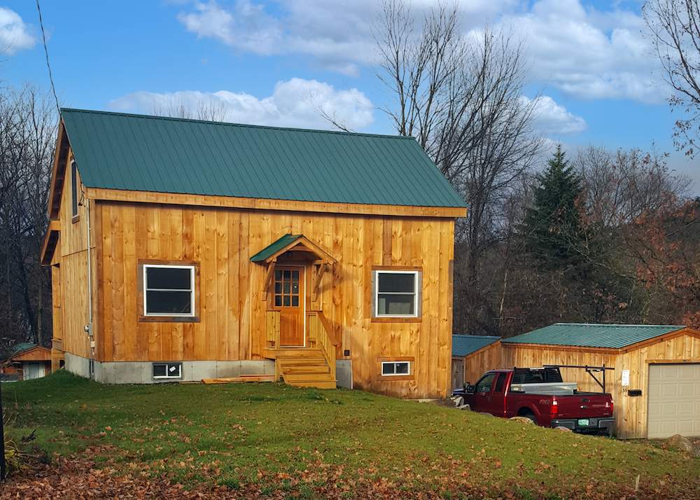 Timber frame cabin with green gable style roof, front door with wood steps and overhang, and grass with fallen leaves.