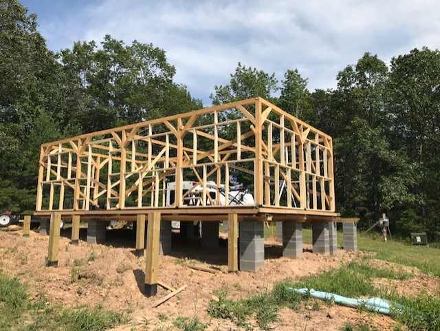 Post and beam timber frame cabin being erected on top of pier foundation on sunny area of grass with trees in the background.