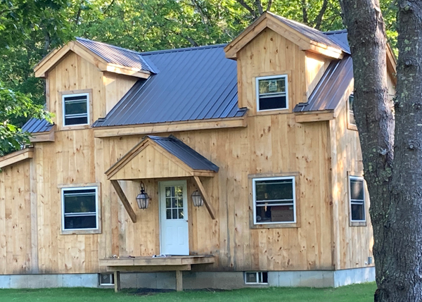 Rustic timber frame post and beam cabin with two doghouse window dormers, a metal roof, and an overhang over the front door.