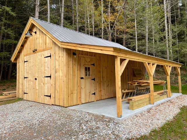 A large post and beam barn with double doors, a gray metal roof, and an overhang in front of a forest scene.