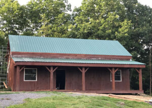 Dark brown post and beam 800-squre-foot small timber frame cabin with green gable style roof, covered porch and green grass.