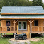 Old man standing on the porch of a rustic cabin with green gable style roof and black ATV parked in front of the porch steps.