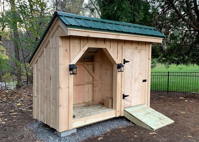 Post and beam wood storage shed with a green metal roof and an open compartment for logs and an enclosed compartment for tools.