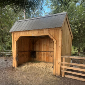 Eight by ten Run-In horse shelter with a gray roof stands next to a fence. There is hay on the ground inside the shed, and behind it is a wooded area.
