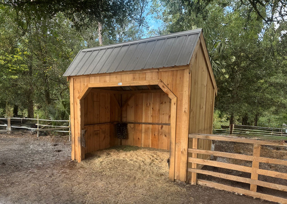 Eight by ten Run-In horse shelter with a gray roof stands next to a fence. There is hay on the ground inside the shed, and behind it is a wooded area.