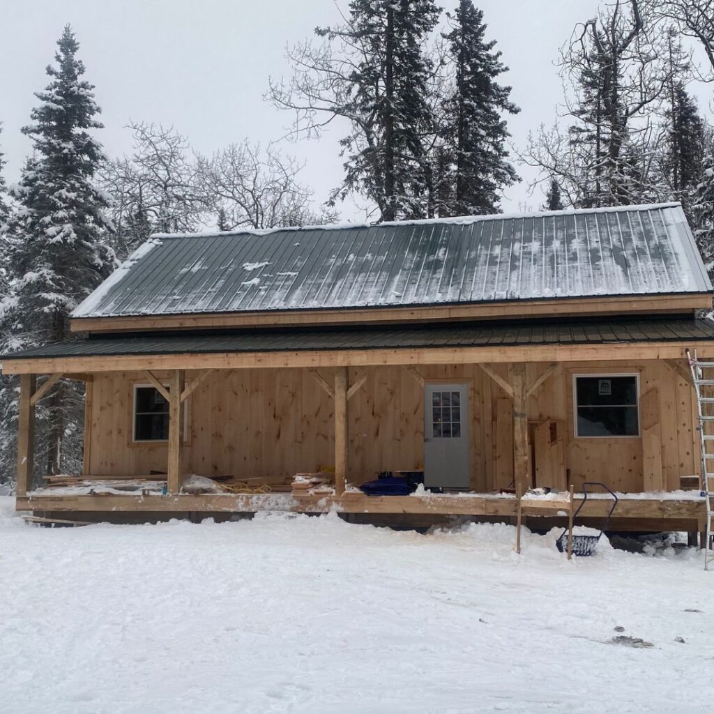 Small rustic cabin in winter with gable style roof and front porch with snowy ground and snowy pine trees behind the cabin.