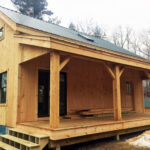 Timber frame post and beam cabin with covered front porch, porch steps, green corrugated metal roof, and snow on the ground.