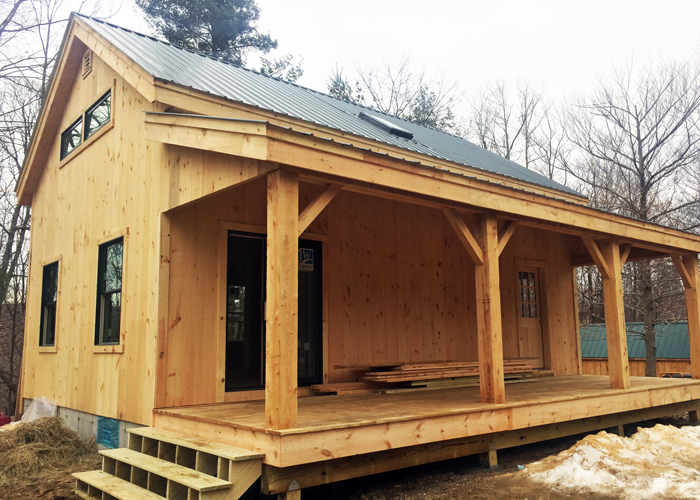 Timber frame post and beam cabin with covered front porch, porch steps, green corrugated metal roof, and snow on the ground.
