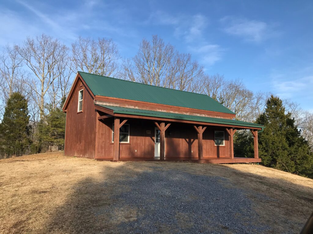 Rustic brown post and beam cabin with green roof and overhang roof over open porch with trees and blue sky behind the home.